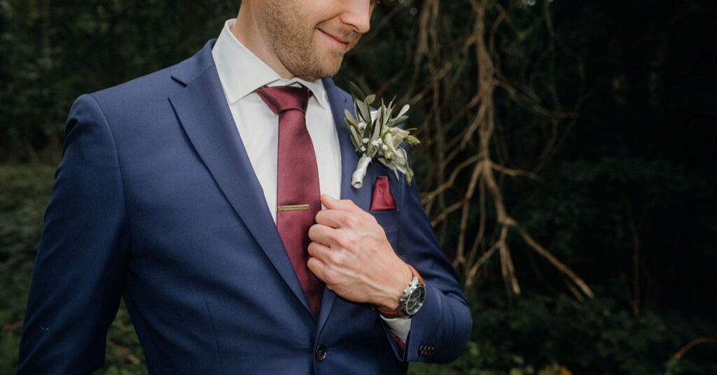 Man wearing a navy blue suit with a burgundy tie and boutonniere.