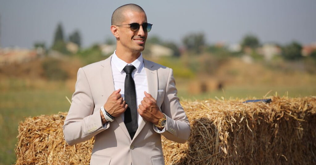 Man wearing a beige suit and black tie outdoors.
