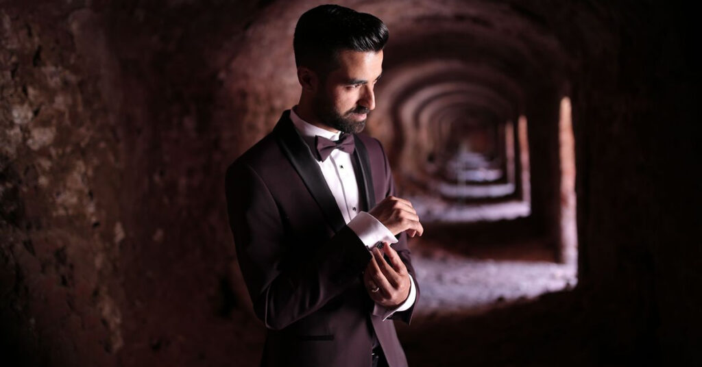 Man wearing a plum coloured suit and bow tie in a formal setting.